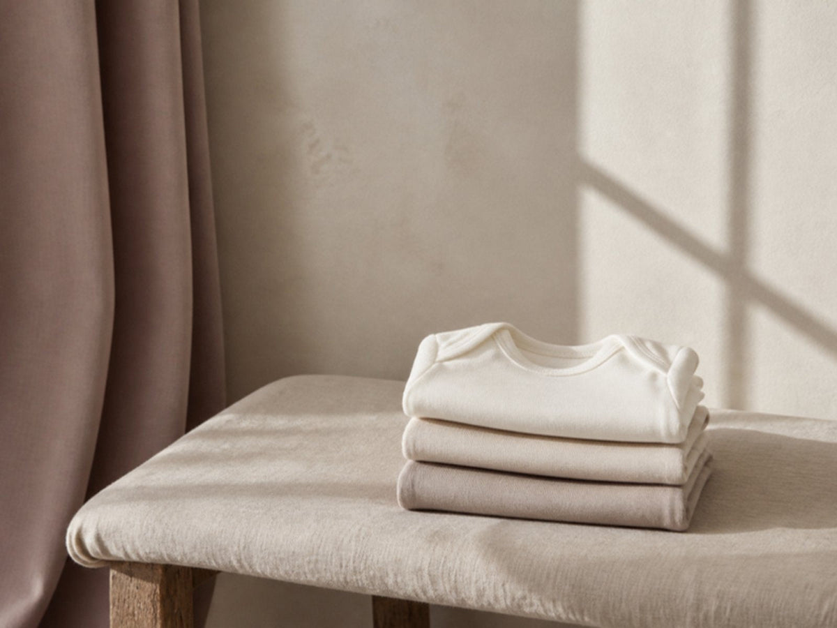 Three folded newborn bodysuits on a linen-covered bench in window light